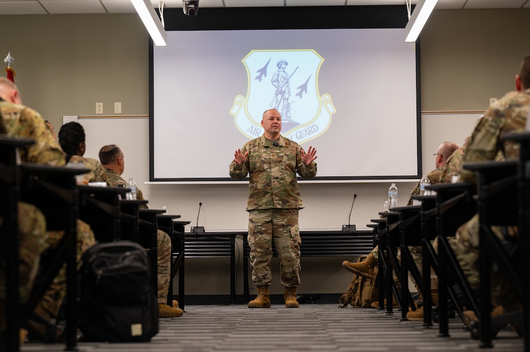 An Airman in an OCP-patterned military is seen standing at the front of a classroom in between rows of uniformed service members. The primary subject is speaking to the group with his hands gesture up at chest-level.