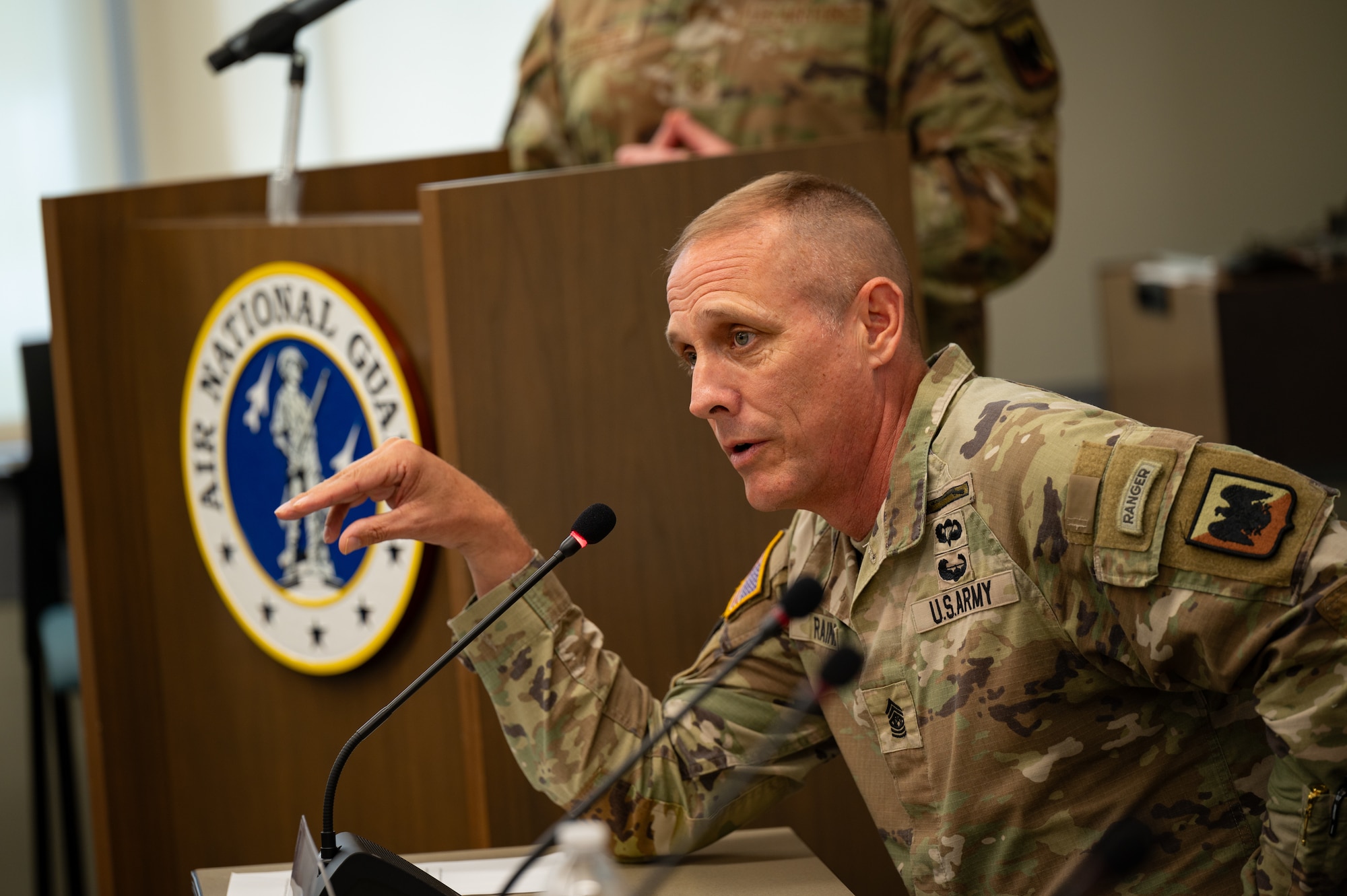 A Solider in an OCP-patterned military uniform leans into a microphone and gestures with his hand while answers a question on a panel.