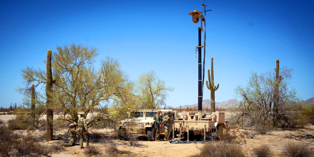 Soldiers gather near a vehicle and a satellite at a makeshift headquarters in a desert-like area.