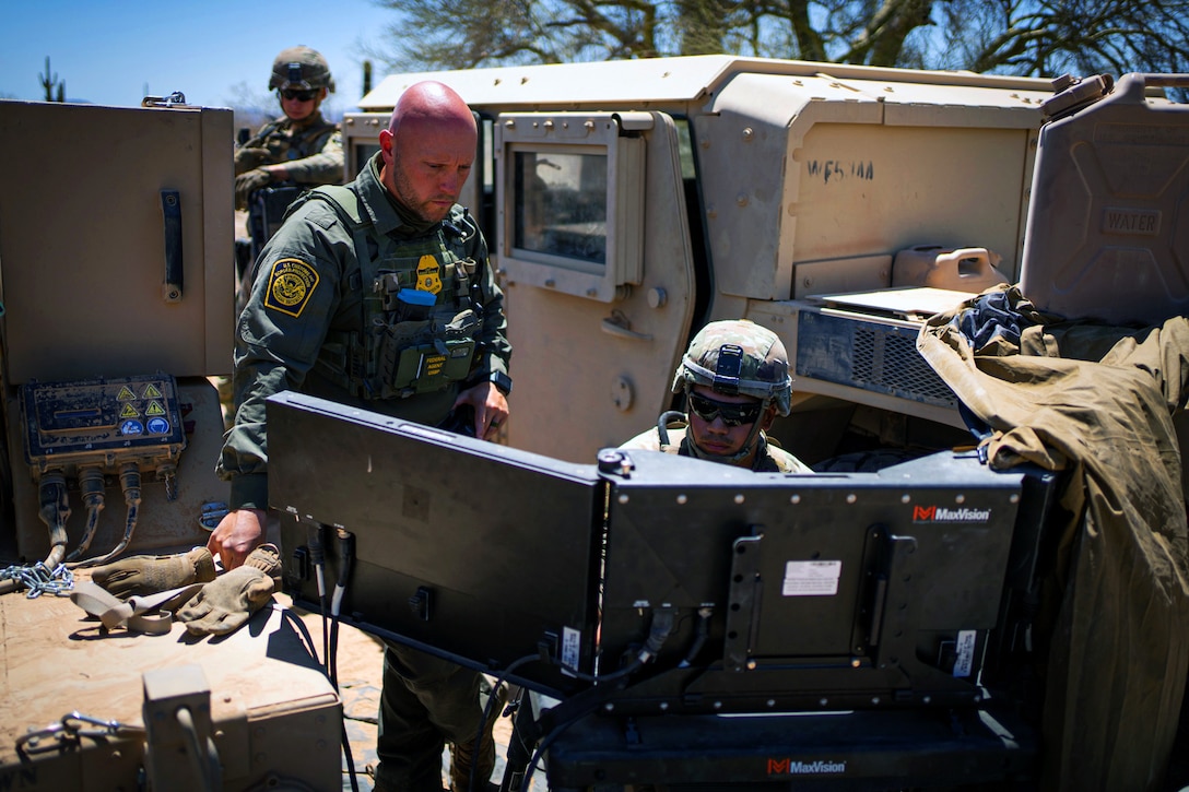 A soldier and a U.S. Border Patrol agent look at monitors during daylight.
