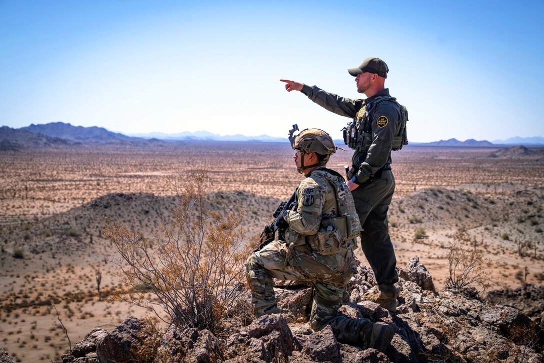 A soldier kneels on rocks as a standing U.S. Border Patrol agent points in the distance in a desert-like area.