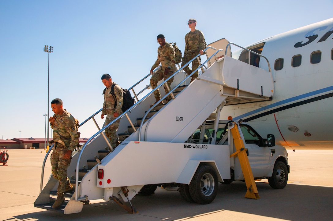 Four soldiers walk down the steps of a parked aircraft on tarmac against a blue sky.