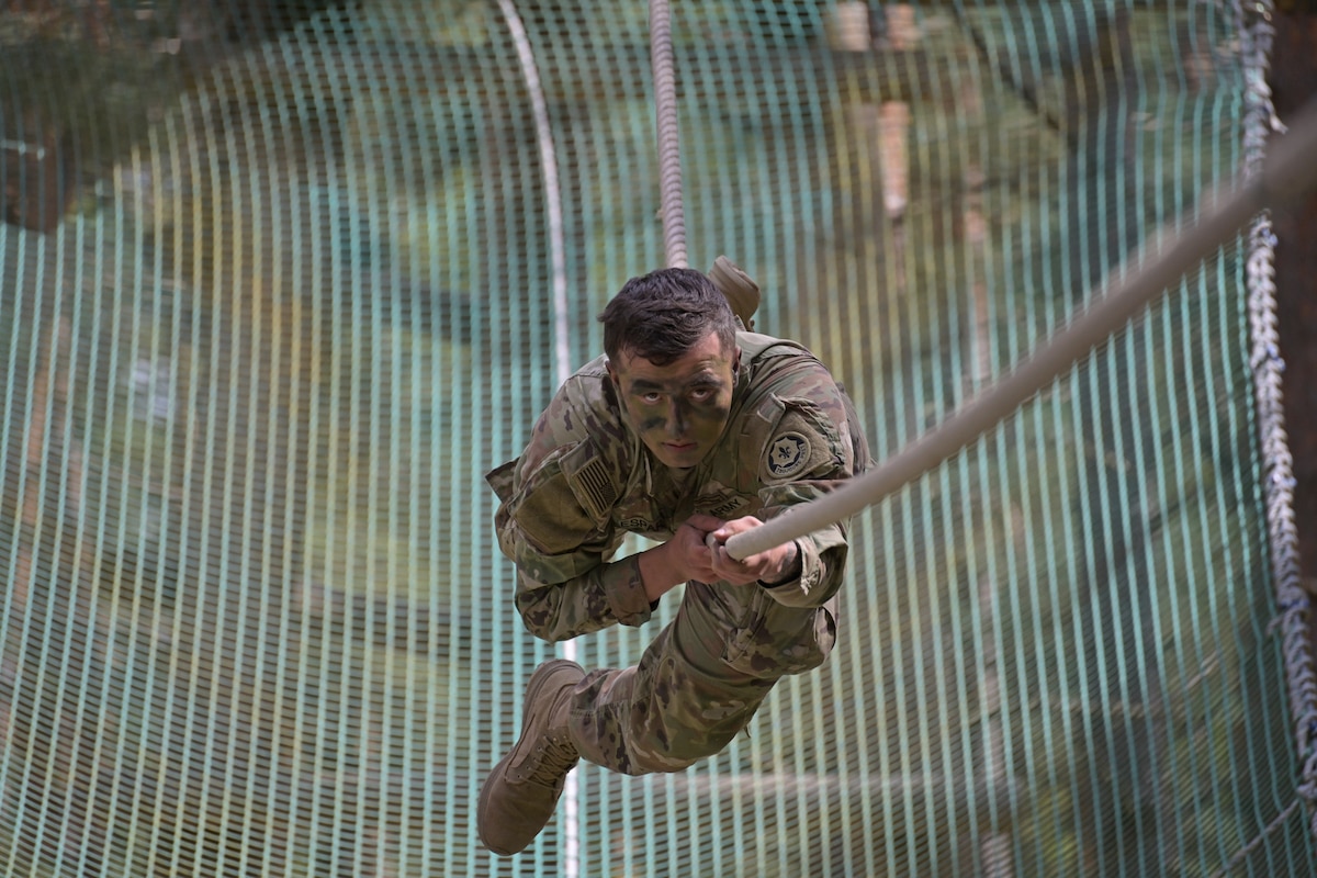 A soldier wearing face paint pulls across a rope obstacle with a net underneath.