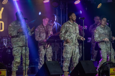 Army band members perform in OCPs on a stage with colorful lights and their microphones.
