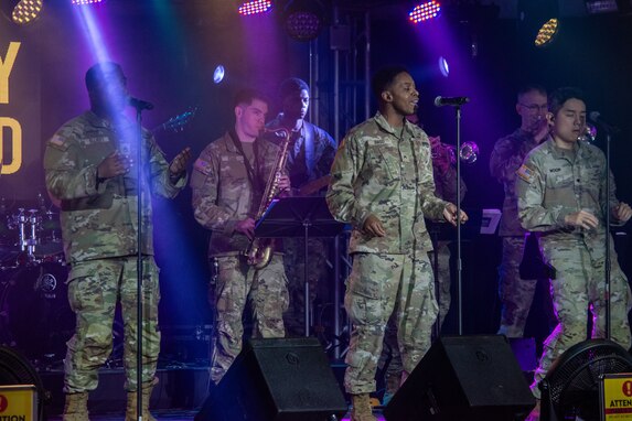 Army band members perform in OCPs on a stage with colorful lights and their microphones.