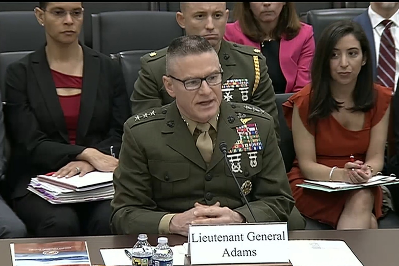 A man in a military uniform speaks into a microphone while a man in a military uniform and several people in business attire sit behind him.
