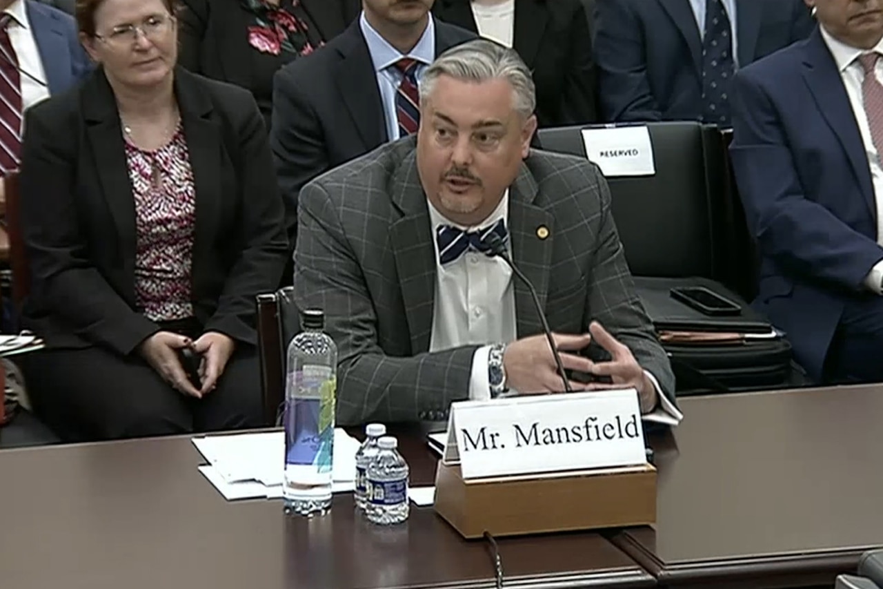 A man in a business suite speaks into a microphone while several people in business attire sit behind him.