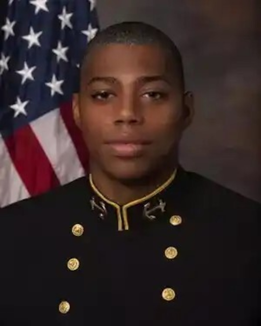 A man wearing a military uniform smiles. An American flag is in the background.