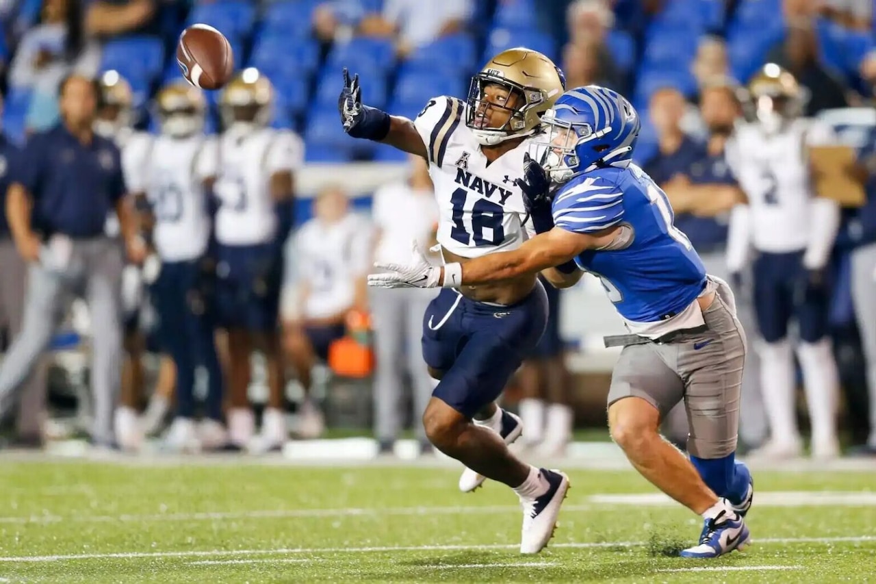 A man wearing a football uniform blocks a football in the air from another man wearing a football uniform.