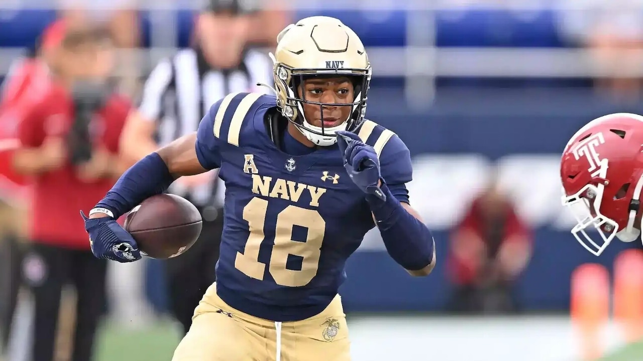 A man wearing a football uniform and carrying a football runs past another man wearing a football uniform.