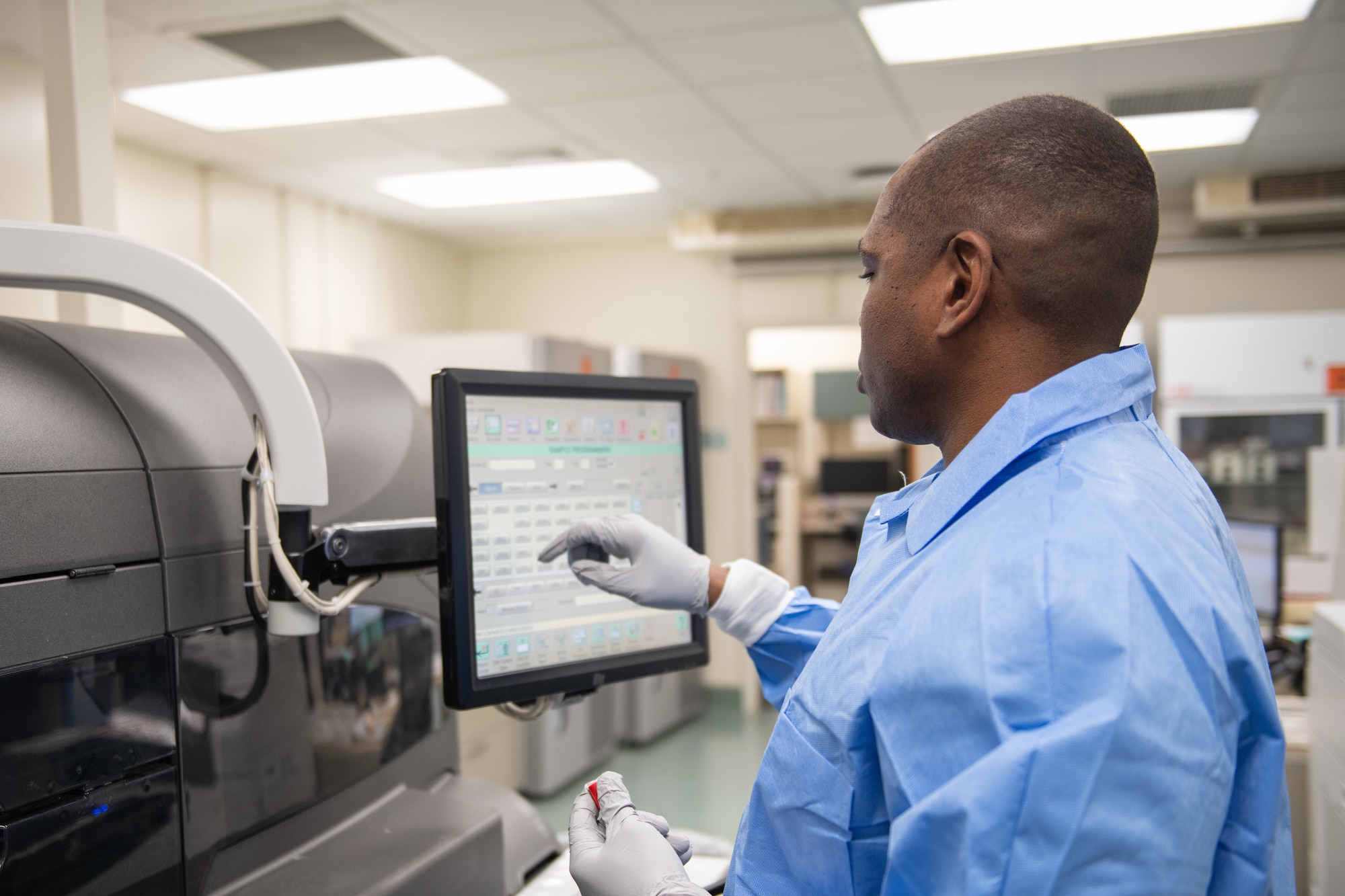 An Airman wearing blue medical scrubs touches a screen attached to a chemistry analyzer.