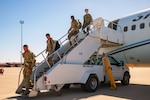 Four soldiers walk down the steps of a parked aircraft on tarmac against a blue sky.
