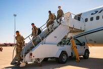 Four soldiers walk down the steps of a parked aircraft on tarmac against a blue sky.