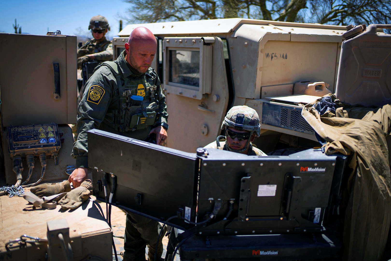 A soldier and a U.S. Border Patrol agent look at monitors during daylight.