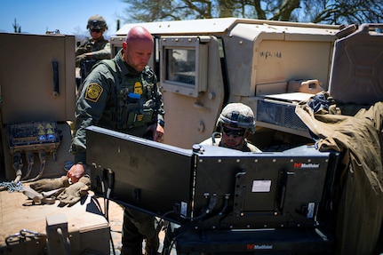 A soldier and a U.S. Border Patrol agent look at monitors during daylight.
