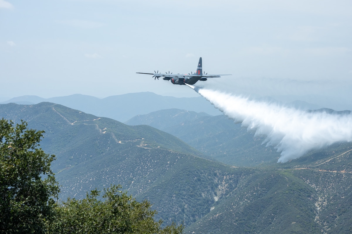 A large military aircraft drops water over a mountainous landscape.