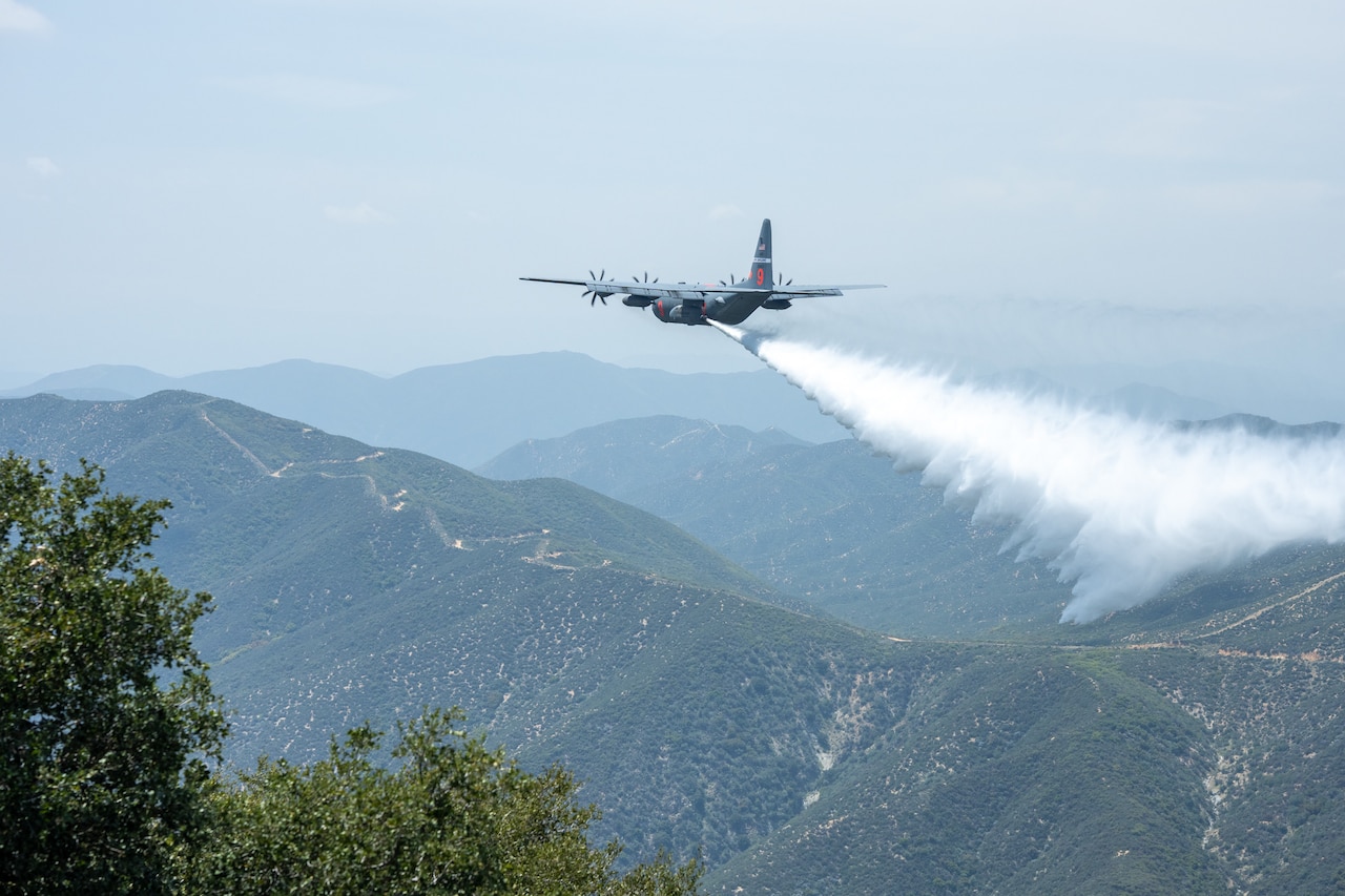 A large military aircraft drops water over a mountainous landscape.