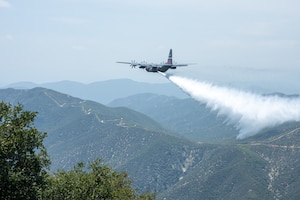 A large military aircraft drops water over a mountainous landscape.