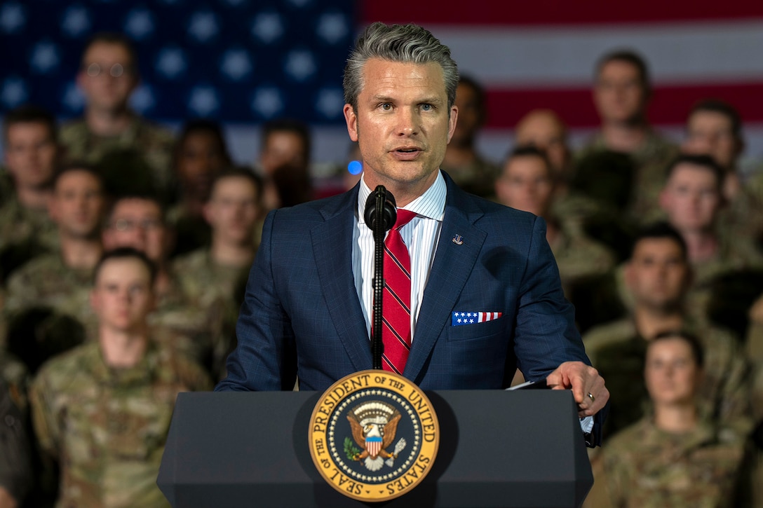 Defense Secretary Pete Hegseth speaks at a lectern with troops standing in the background.
