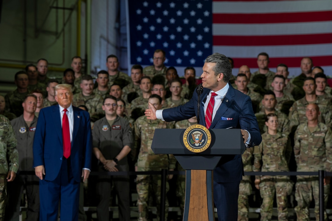 Defense Secretary Pete Hegseth gestures from a lectern toward President Donald J. Trump, who stands with troops.