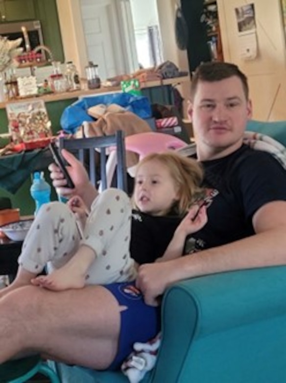 Man and young daughter sit in a sofa chair in their house.