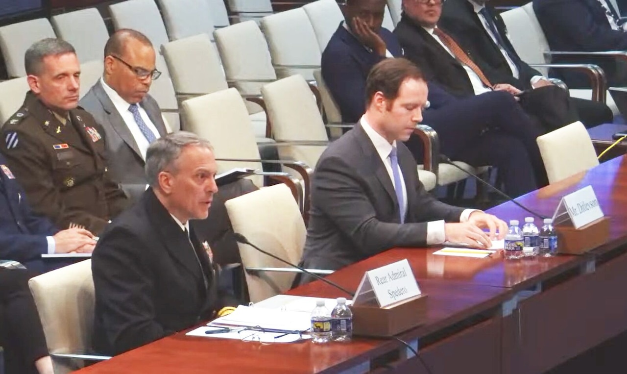 Two men, one in a military uniform and the other in business attire, sit at a long table. Several men in military uniforms and business attire sit behind them.