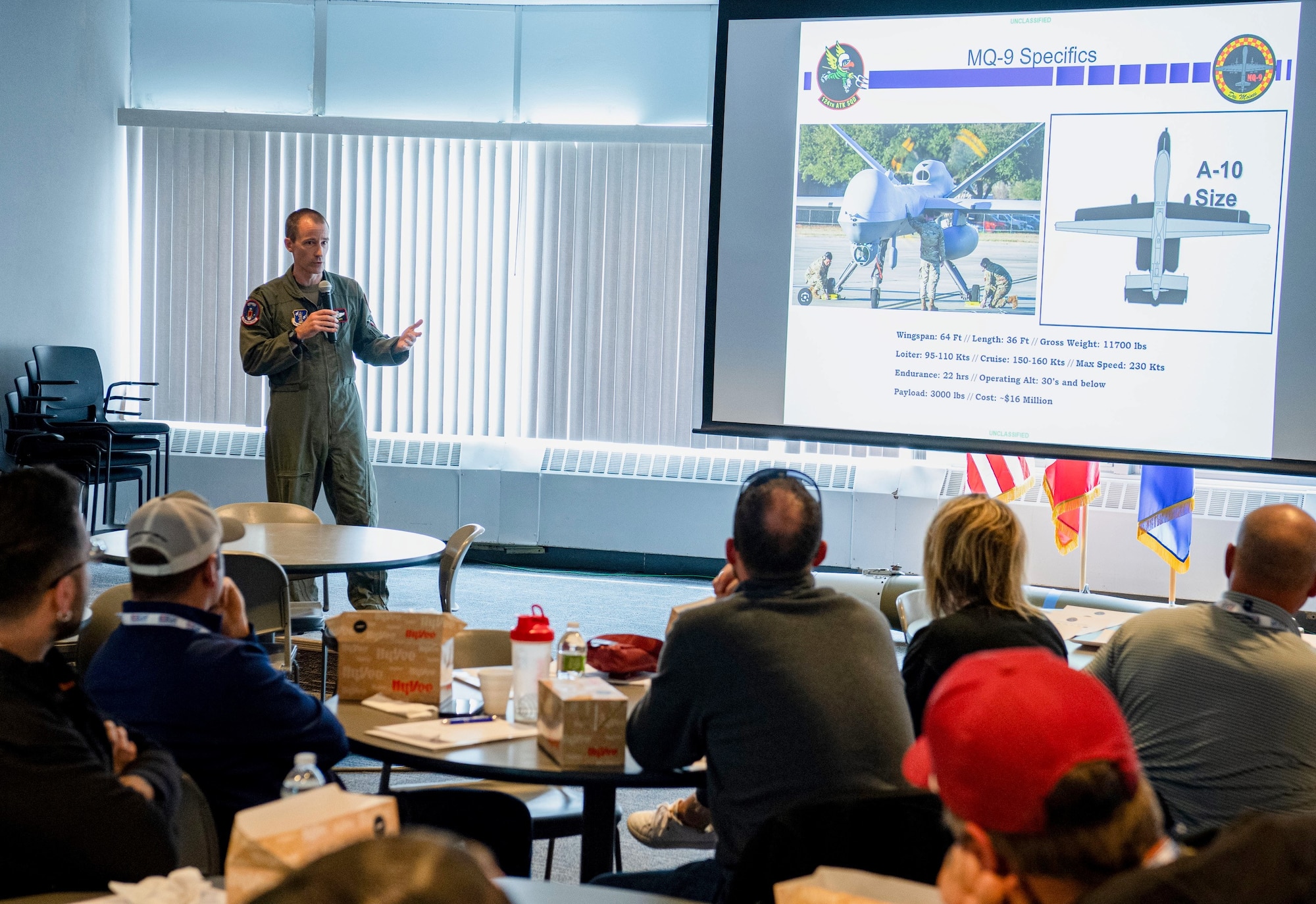 Air Force pilot in green flight suit talks to group of people by a screen.