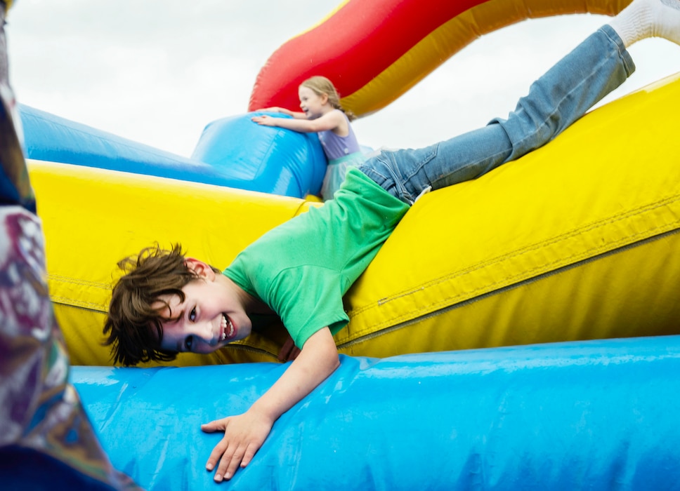 A boy, in a green tee shirt, slides over the obstacles of a bounce house laying on his stomach.