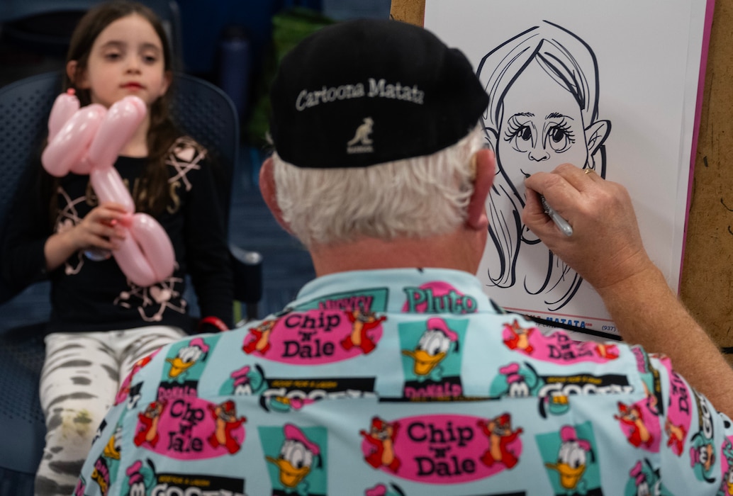 A small girl, holding a balloon animal, poses as a man, whose back is to the camera, sketches her.