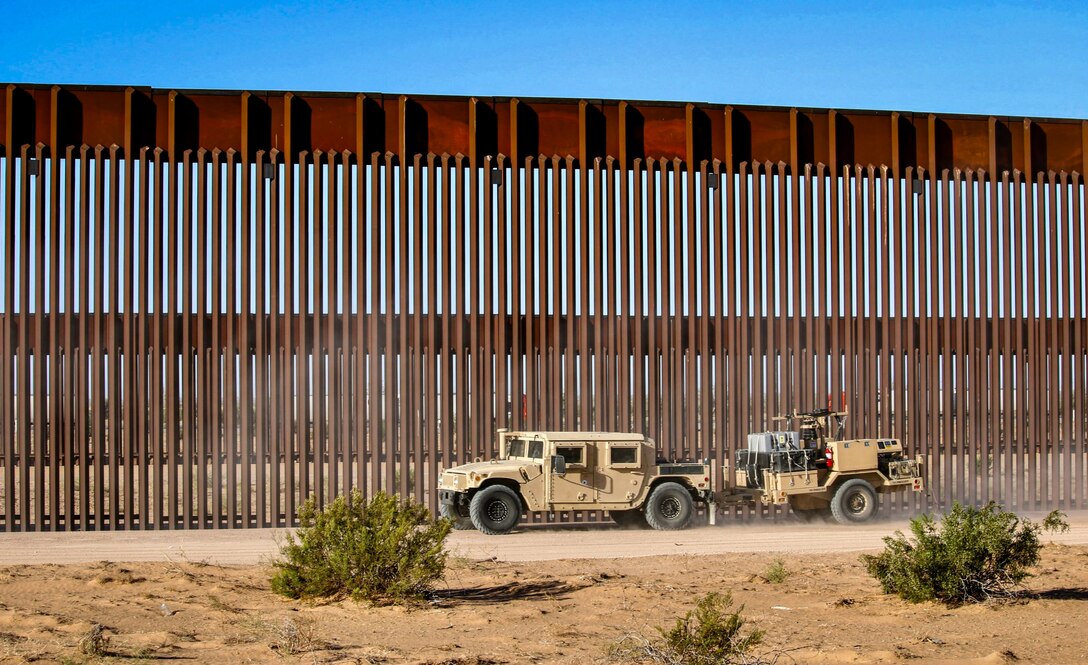 Soldiers drive vechiles near a large outdoor wall in a desert-like area against a blue sky.