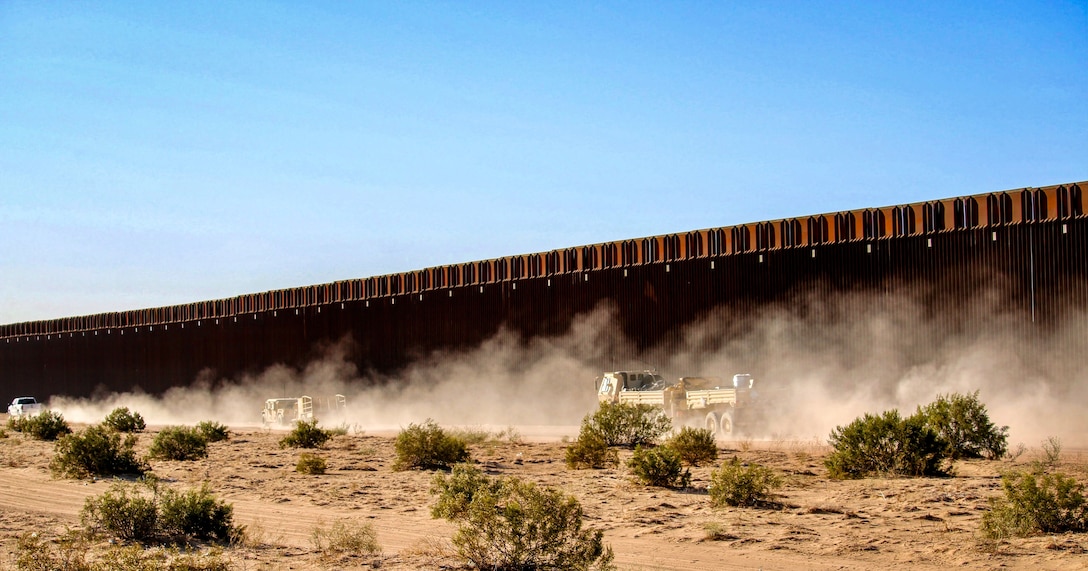Dust blows in the air as vehicles drive on a dirt road near a large outdoor wall in a desert-like area against a blue sky.