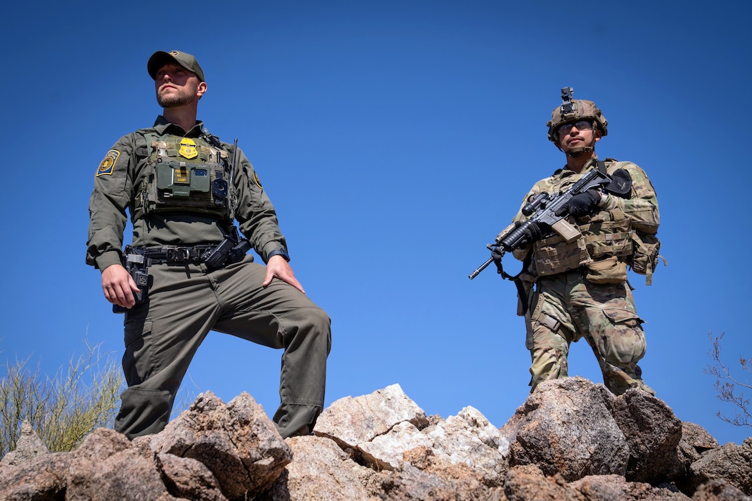A soldier carrying a weapon and a U.S. Border Patrol agent stand on rocks against a blue sky.