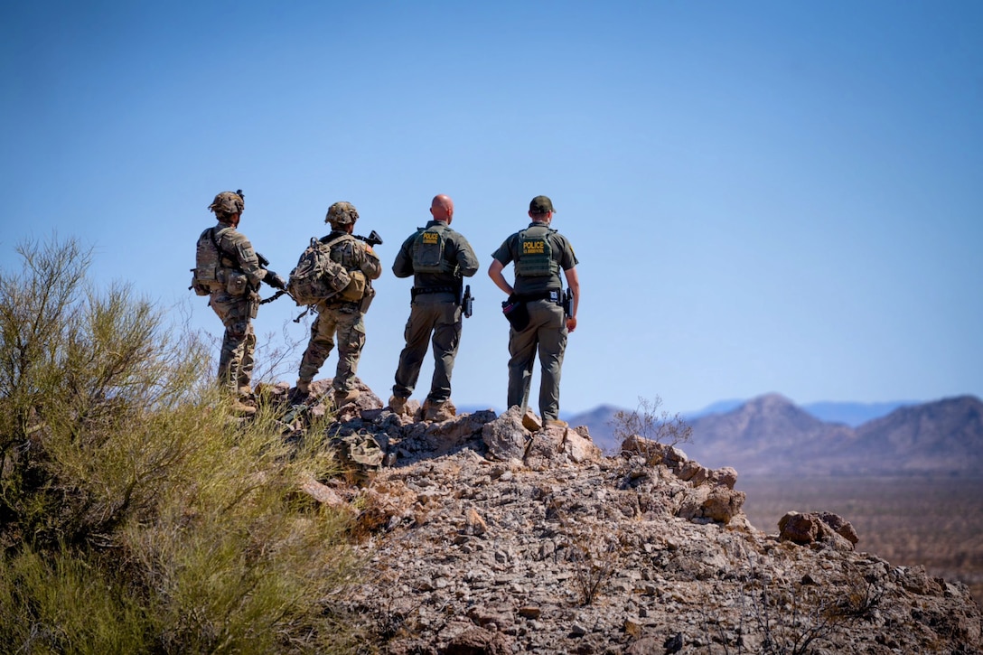 Two soldiers and two U.S. Border Patrol agents standing on rocky terrain look toward mountains.
