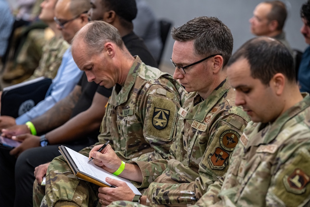 A group of service members in uniform and civilians in business casual take notes