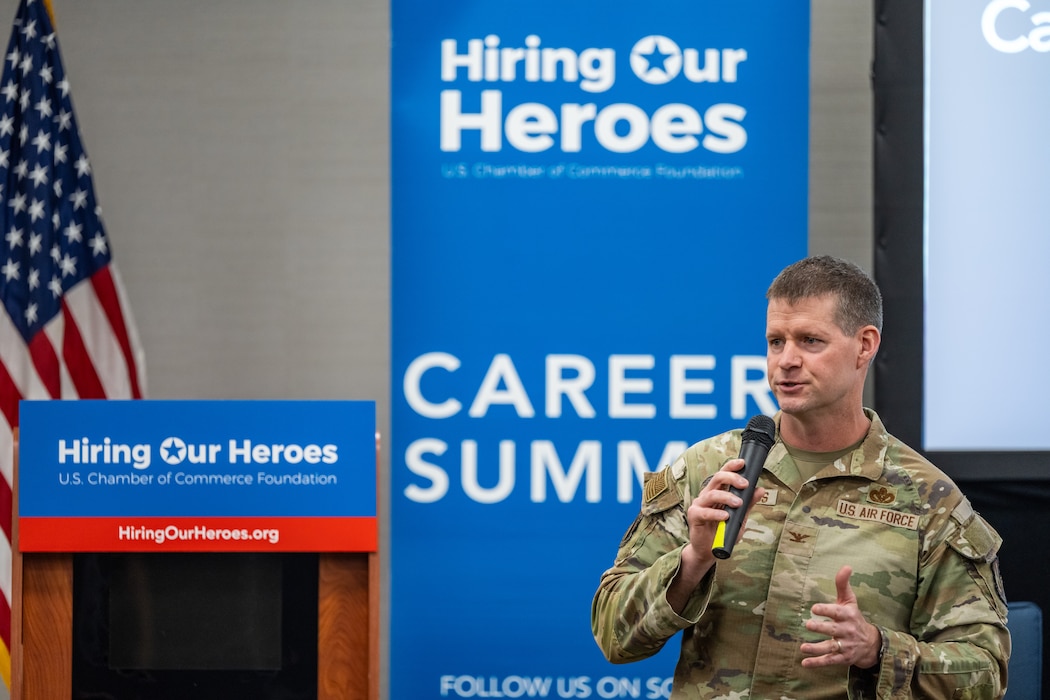 Col. Richards, in military uniform, holds a microphone while speaking in front of a Hiring Our Heroes banner and U.S. flag