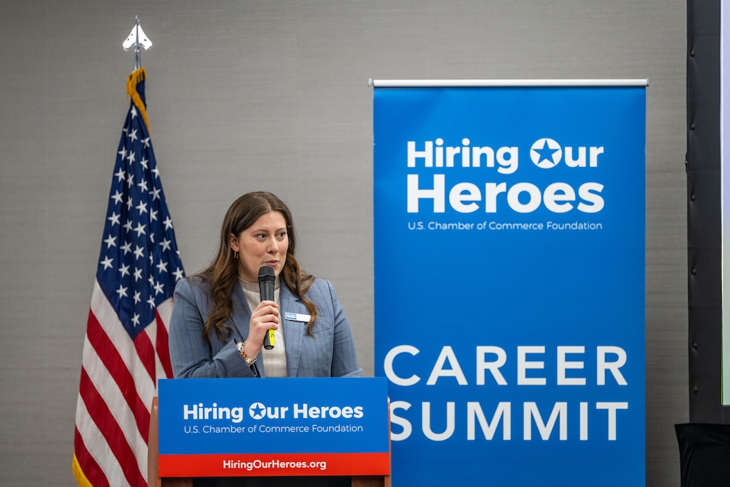 A woman in a bluish grey blazer holds a microphone as she speaks behind a podium with Hiring Our Heroes banners and U.S. flag behind her