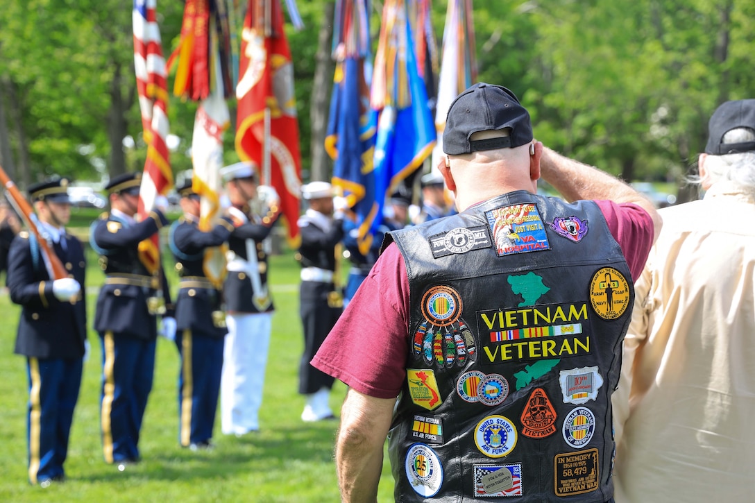A veteran in a leather vest with patches salutes service members next to standing flags outside.