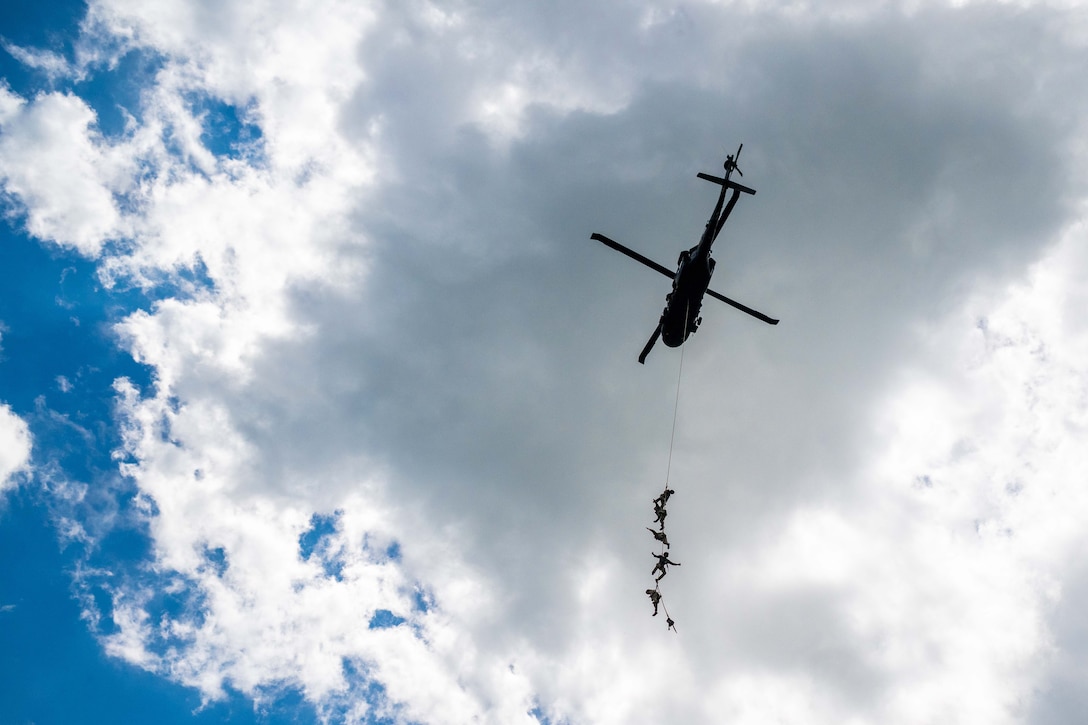 Five soldiers harnessed to a long rope hang from a military helicopter in flight.