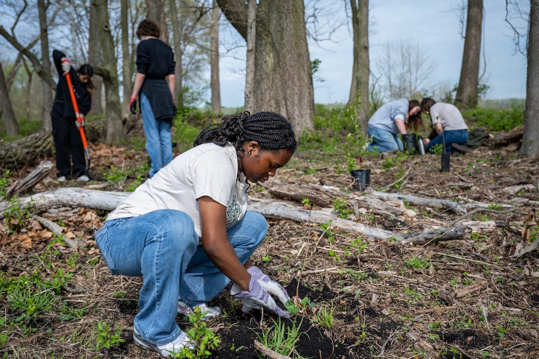 A young high school student bends down to plant a new tree in the dirt, other students can be seen in the background