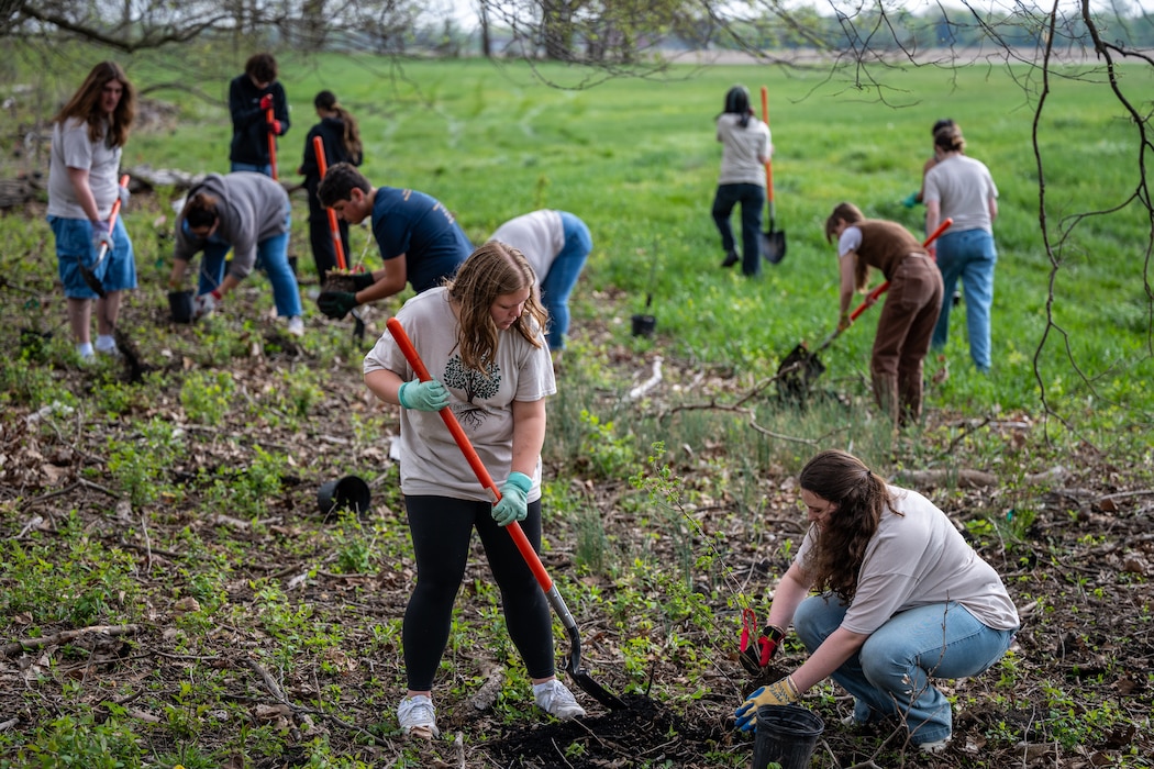 A group of high school students with shovels and potted plants are scattered though the image