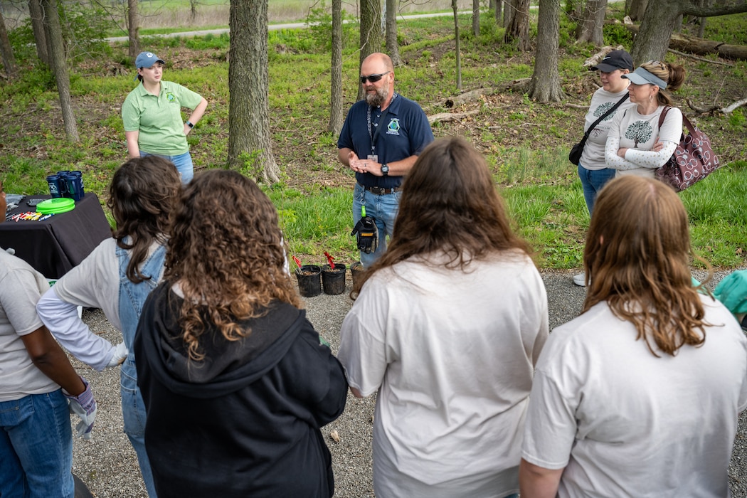 A group of people stand in a semi-circle outdoors among trees as a man in a blue polo and sunglasses speaks in the middle