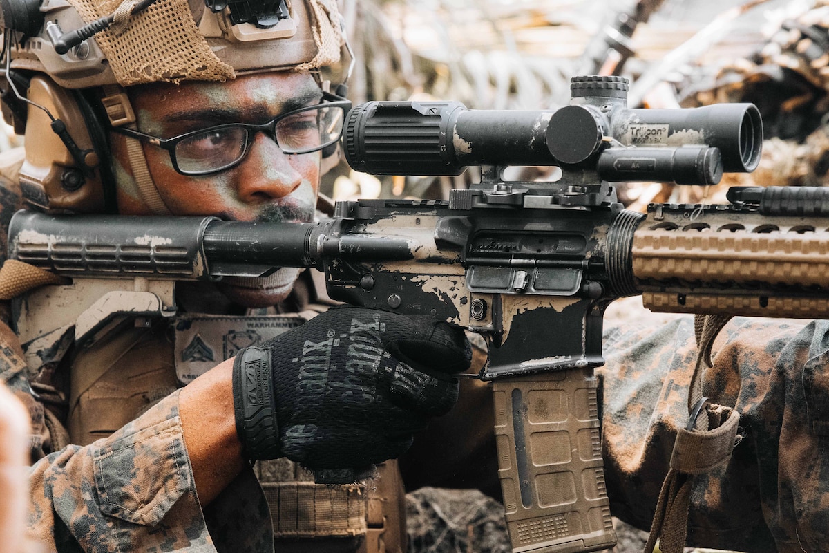 A Marine in tactical gear aims a weapon and looks through its scope.