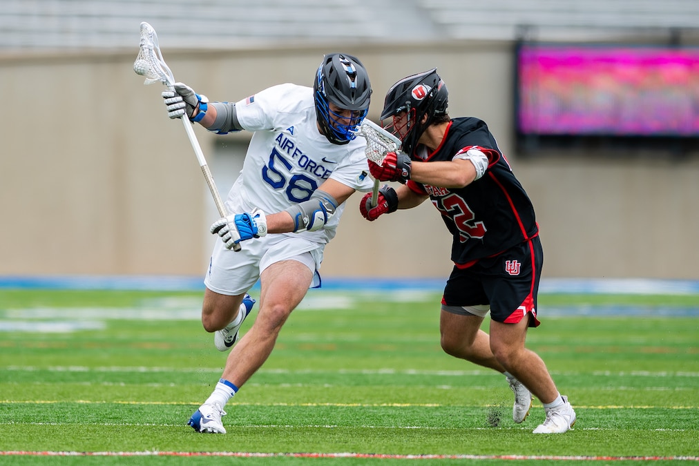 An athlete in white runs into an athlete in black while both hold lacrosse sticks on a green field.