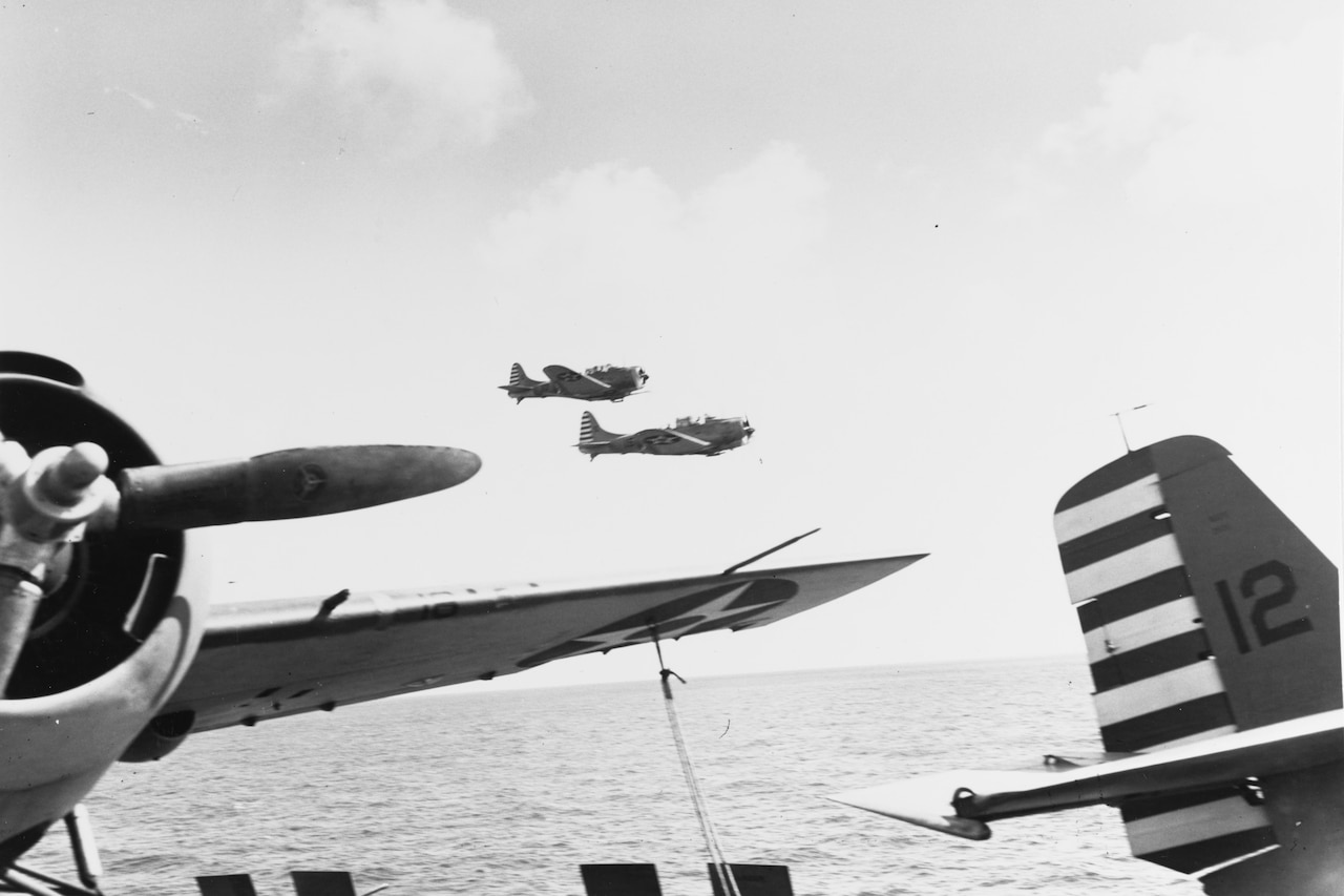 Two aircraft fly low over water near an aircraft carrier flight deck where two other aircraft after perched.