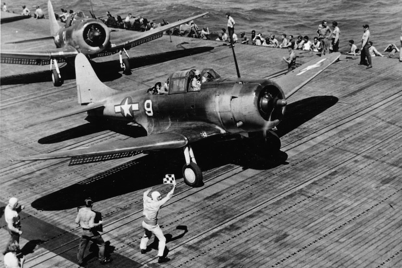 A man waves a flag at a propeller aircraft on an aircraft carrier flight deck.
