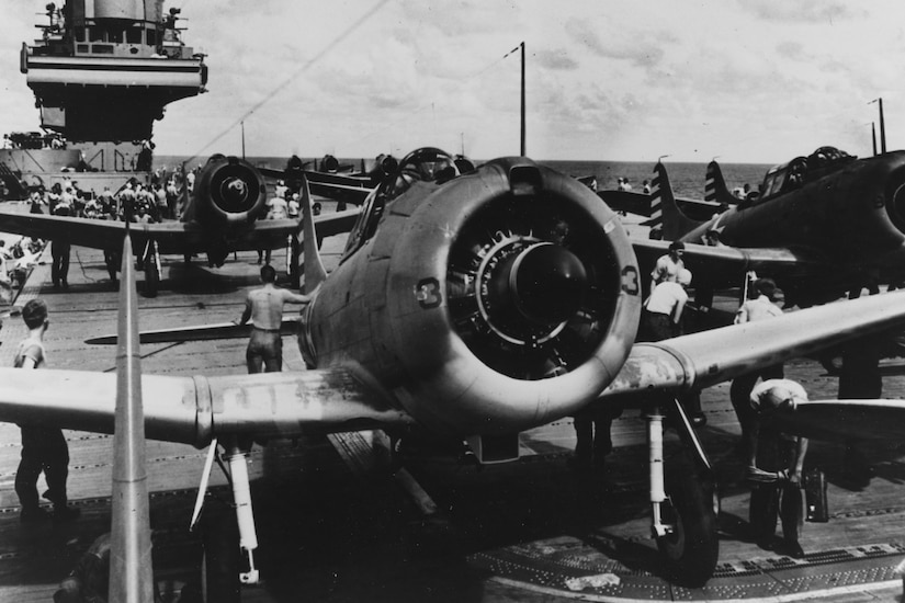 Several propeller aircraft sit on an aircraft carrier's tarmac while sailors reposition them.