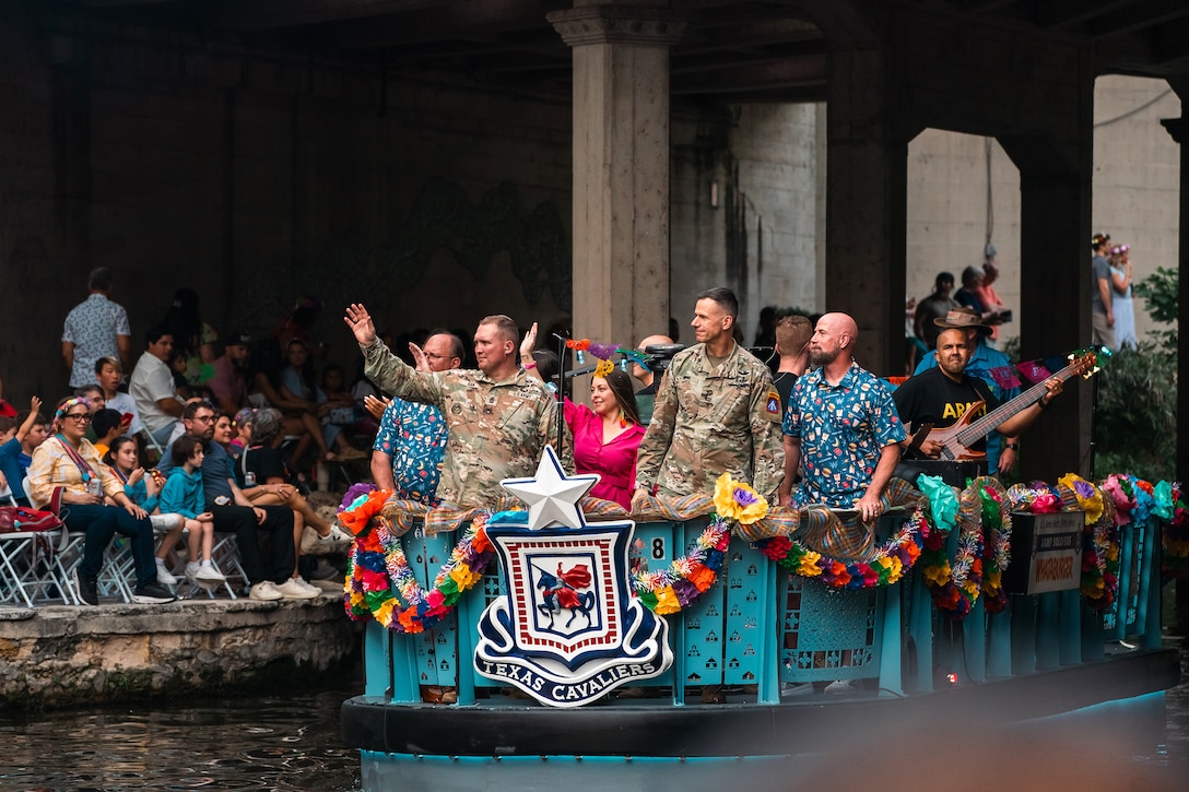 Two soldiers stand with a waving crowd on a float on a waterway looking at people seated on shore.