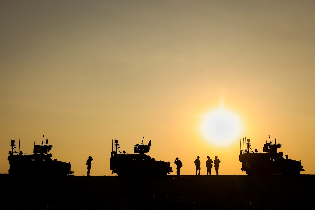 A low sun in an orange sky creates a silhouetted view of Marines standing between three vehicles.