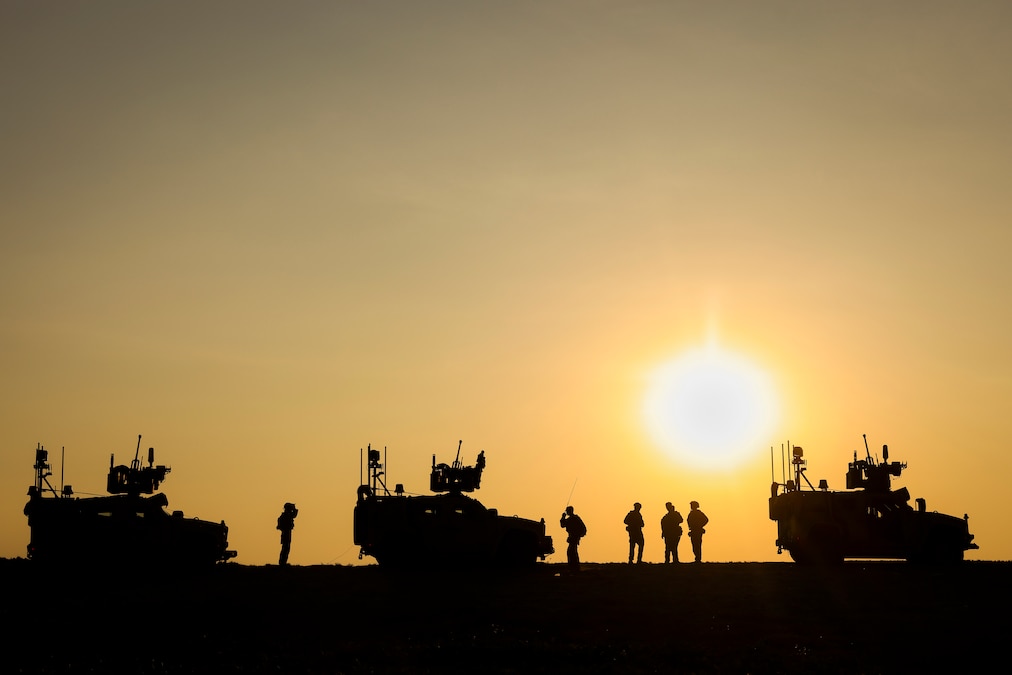 A low sun in an orange sky creates a silhouetted view of Marines standing between three vehicles.