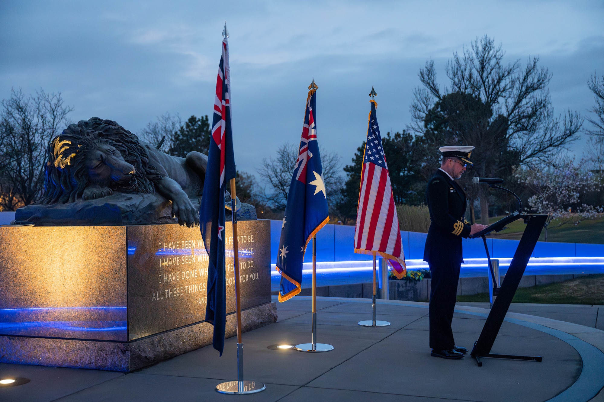 Royal Australian Navy Cmdr. Graeme Davidson stands in front of a lectern with the U.S., Australian and New Zealand flags behind him.